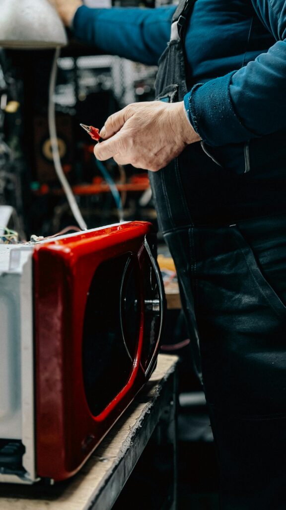 Electrician working on red microwave repair in a workshop setting, tools in hand.
